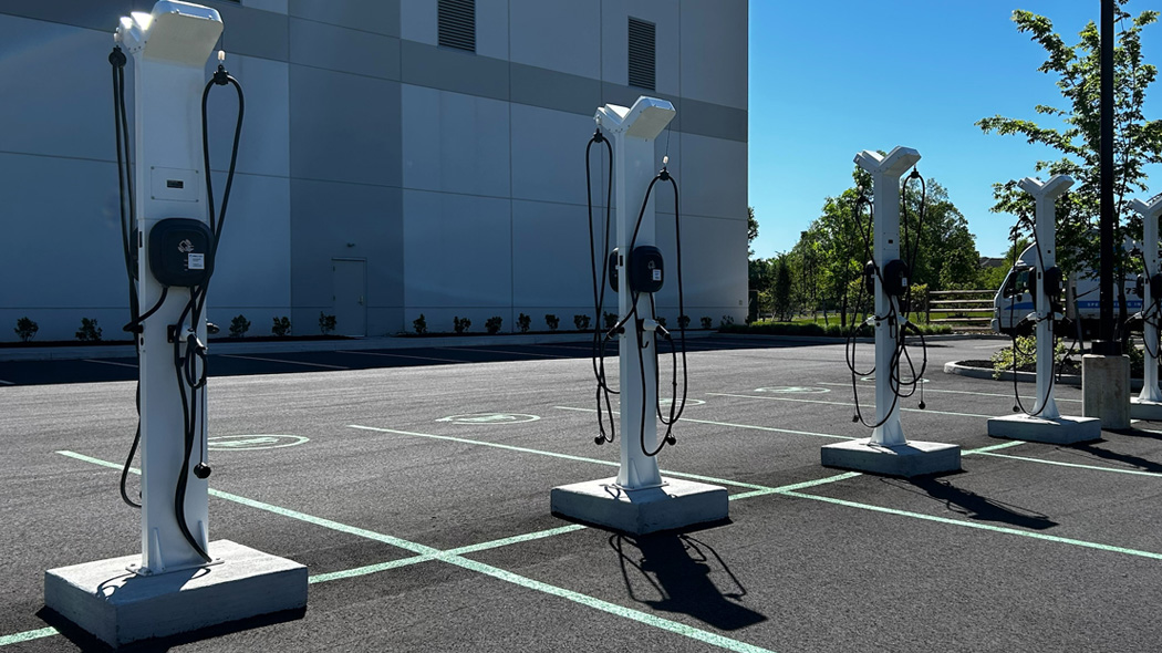 Ground view of 4 EV-charging stations on a sunny day with industrial building behind them in parking lot